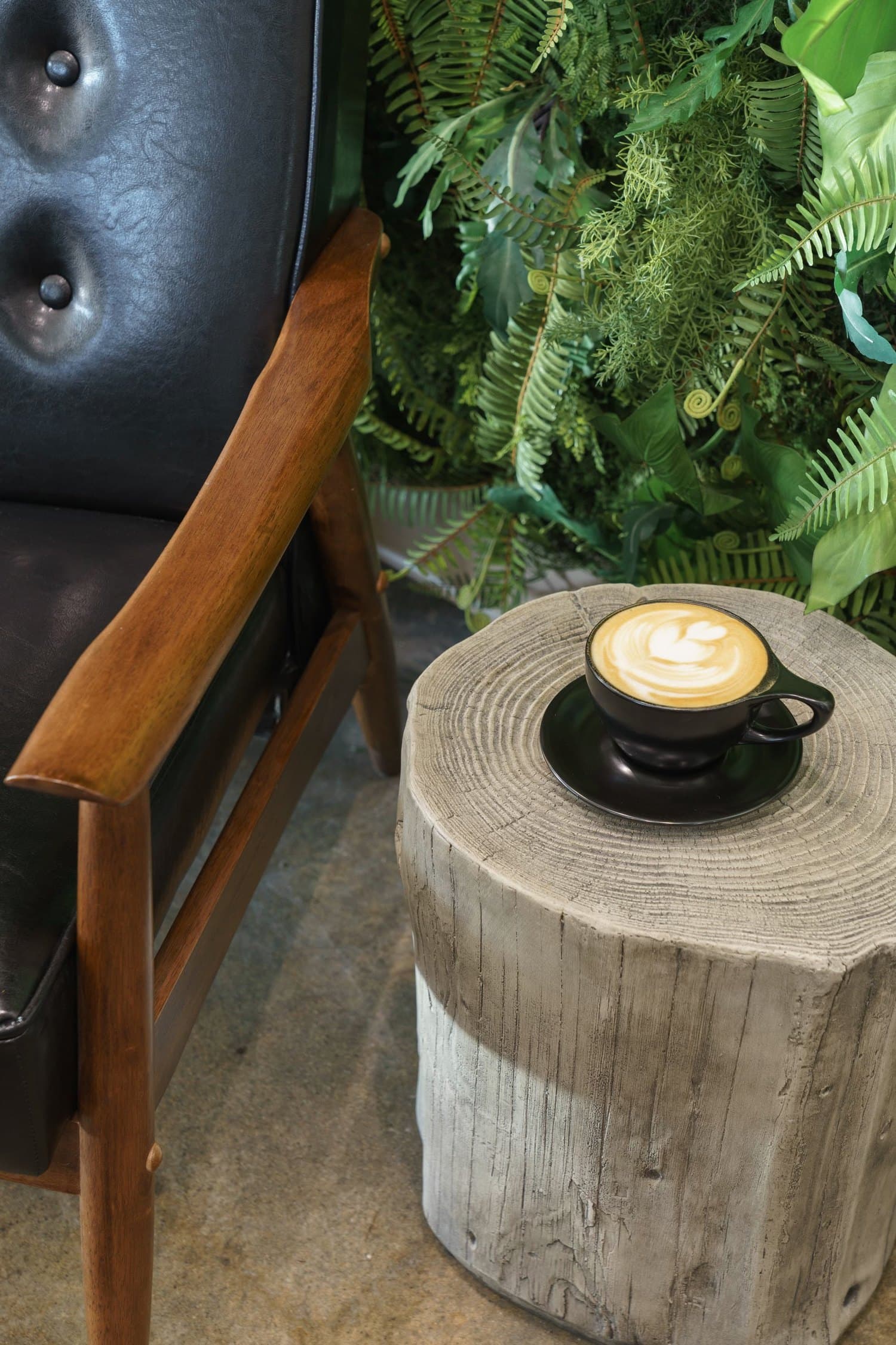 A latte on a wood stump table beside a leather chair and ferns