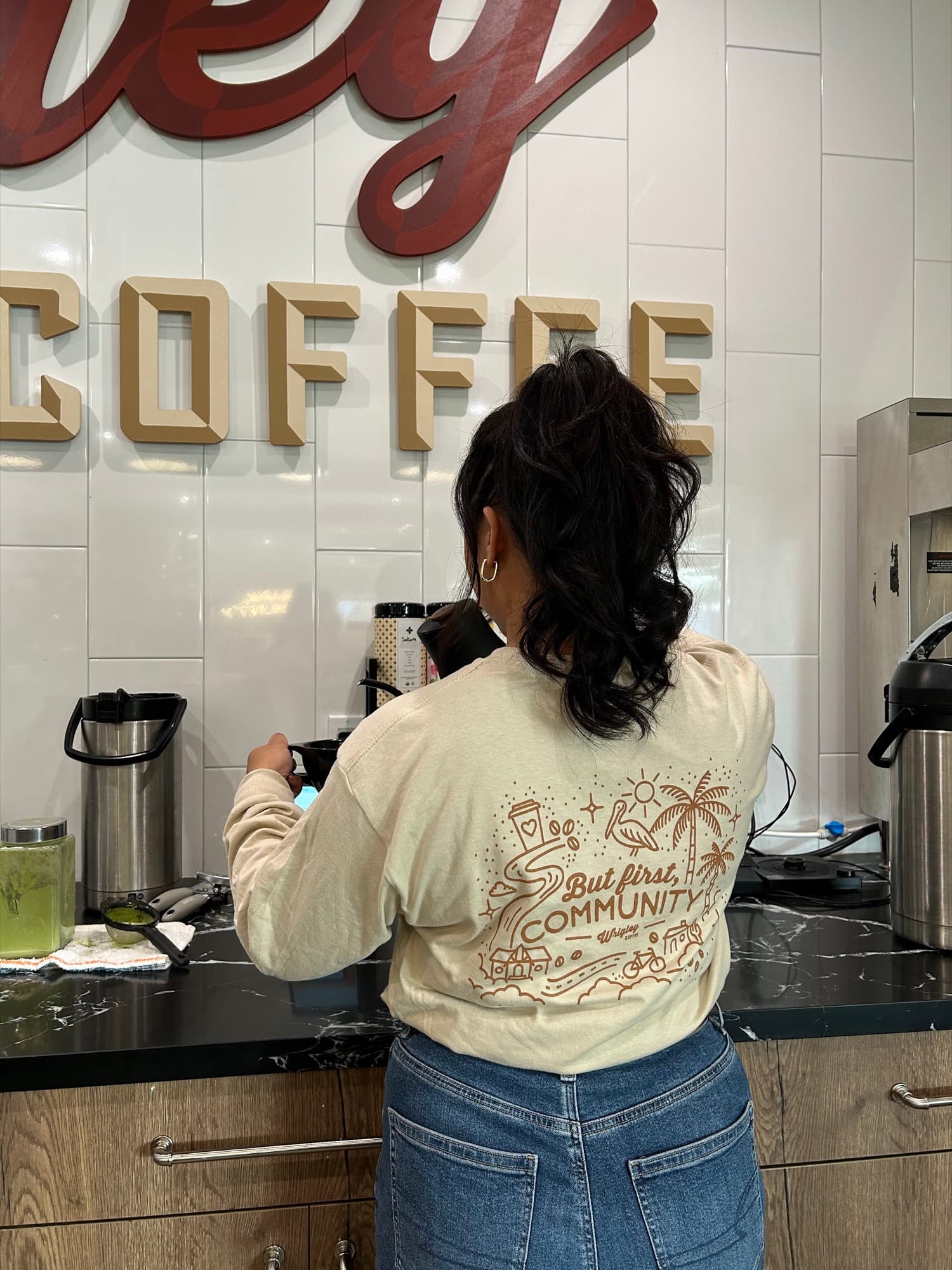 A Wrigley Coffee team member at the coffee station, wearing a sweatshirt that reads But first, Community