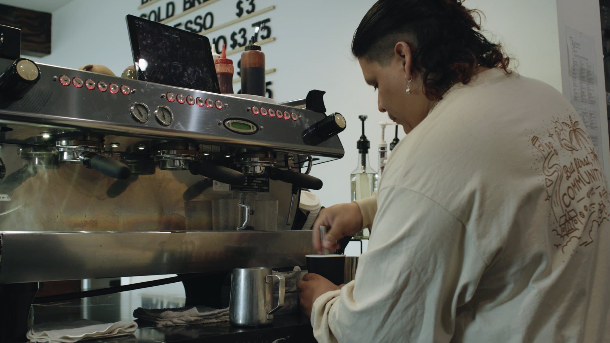 A Wrigley Coffee team member makes a drink at the espresso machine, wearing a shirt that reads But first, Community