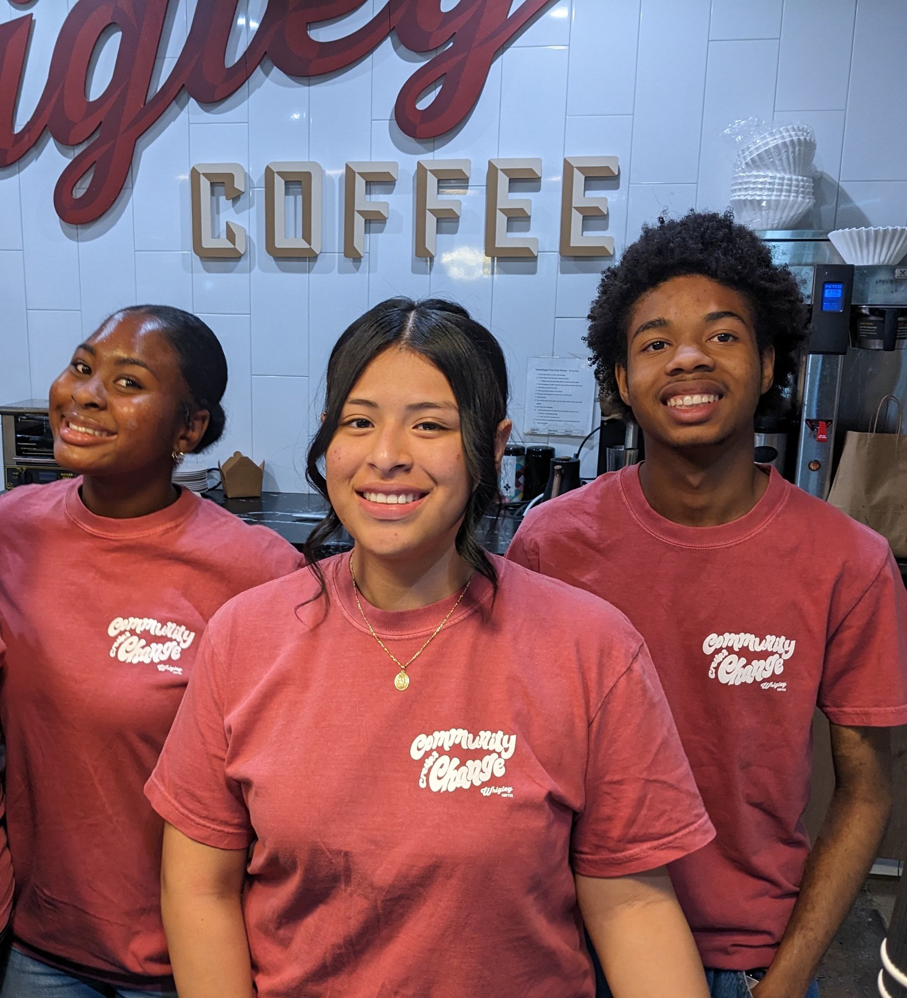 Three spring 2026 pilot students in red shirts smile behind the counter, under the Wrigley Coffee sign